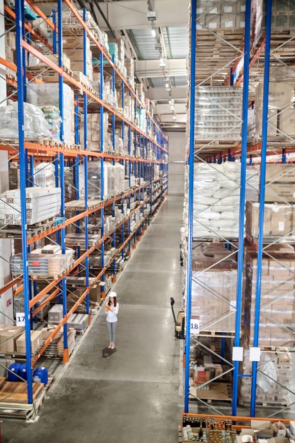 Large warehouse. Woman in white protective helmet with tablet standing in large industrial warehouse in aisle between high racks for goods